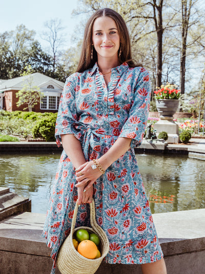 Woman standing near a garden fountain, wearing a teal floral ruffle-collar dress with a tie waist and 3/4 sleeves. She holds a woven basket filled with fresh citrus fruit and accessorizes with gold jewelry and statement earrings.