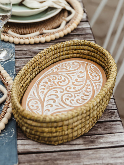 Bread warmer set on outdoor wooden dining table. Ceramic warmer has swirling design and is place inside a handwoven basket. 