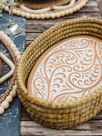 Bread warmer set on outdoor wooden dining table. Ceramic warmer has swirling design and is place inside a handwoven basket. 