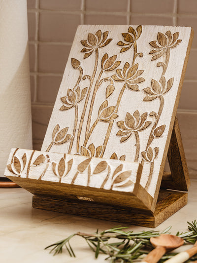 Wooden cookbook holder with lotus flower deign placed on counter top with greenery in the corner. 