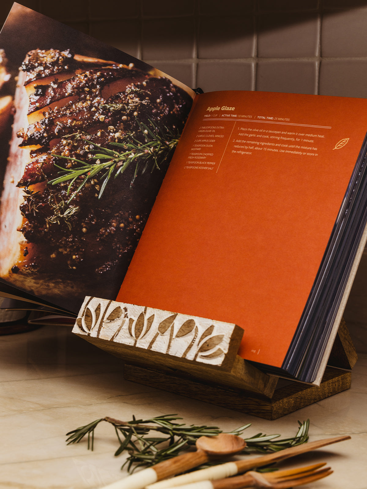 Wooden cookbook holder with lotus flower deign placed on counter top with book placed in holder and  with greenery in the corner. 