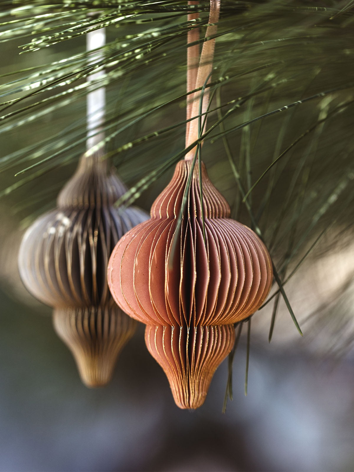 Two handcrafted peach and grey paper ornaments with intricate folds, hanging from a pine tree branch, creating a natural and festive holiday atmosphere.