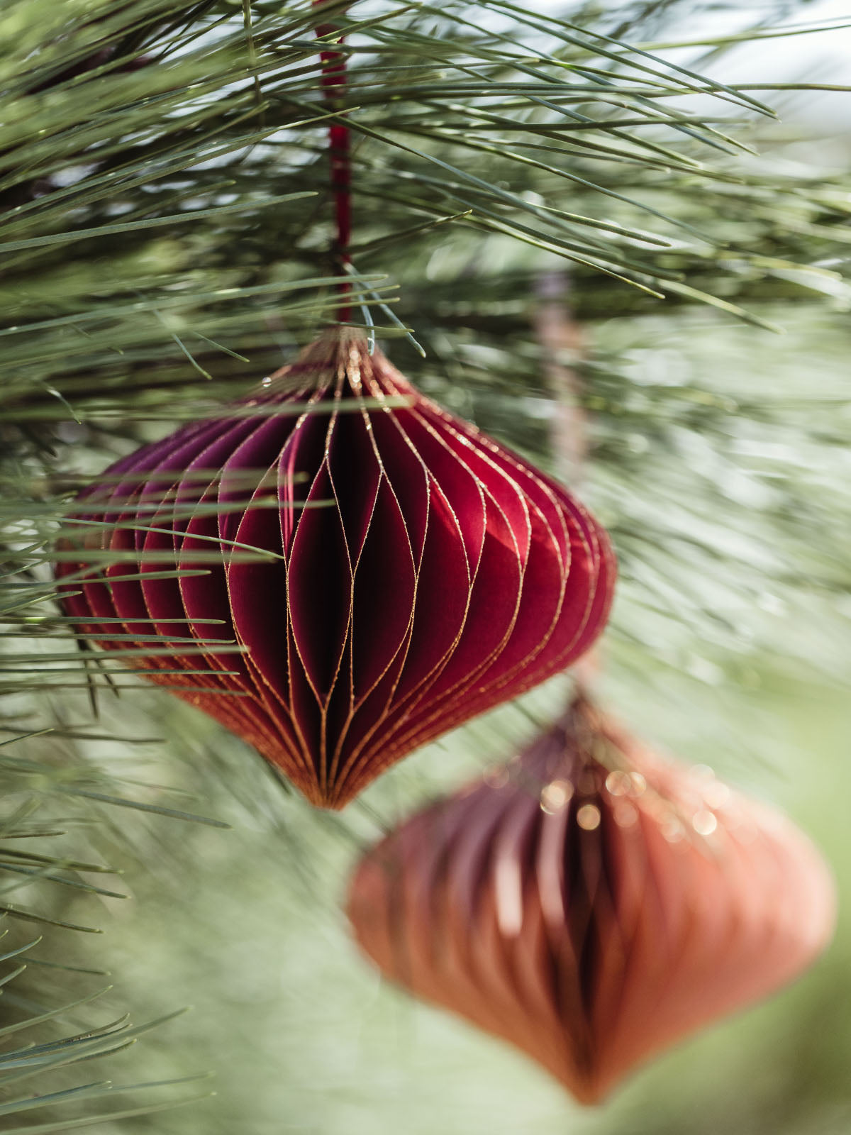 Red and peach handcrafted paper ornaments with gold accents, hanging from a pine tree branch, adding a rich, elegant touch to holiday decor.