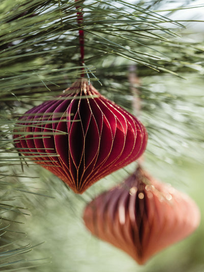 Red and peach handcrafted paper ornaments with gold accents, hanging from a pine tree branch, adding a rich, elegant touch to holiday decor.