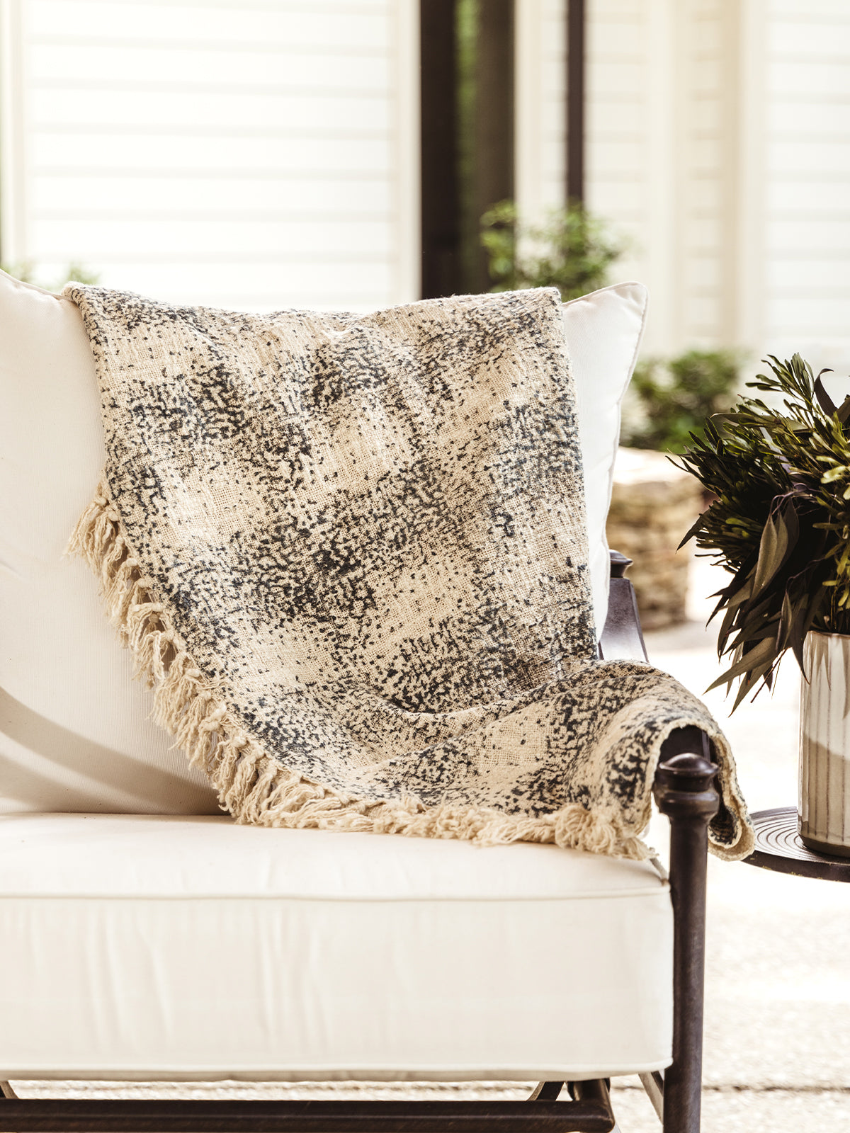 Dusk to Dawn throw blanket on outdoor patio chair. Blanket is cream and spelled with a grey-blue color. 