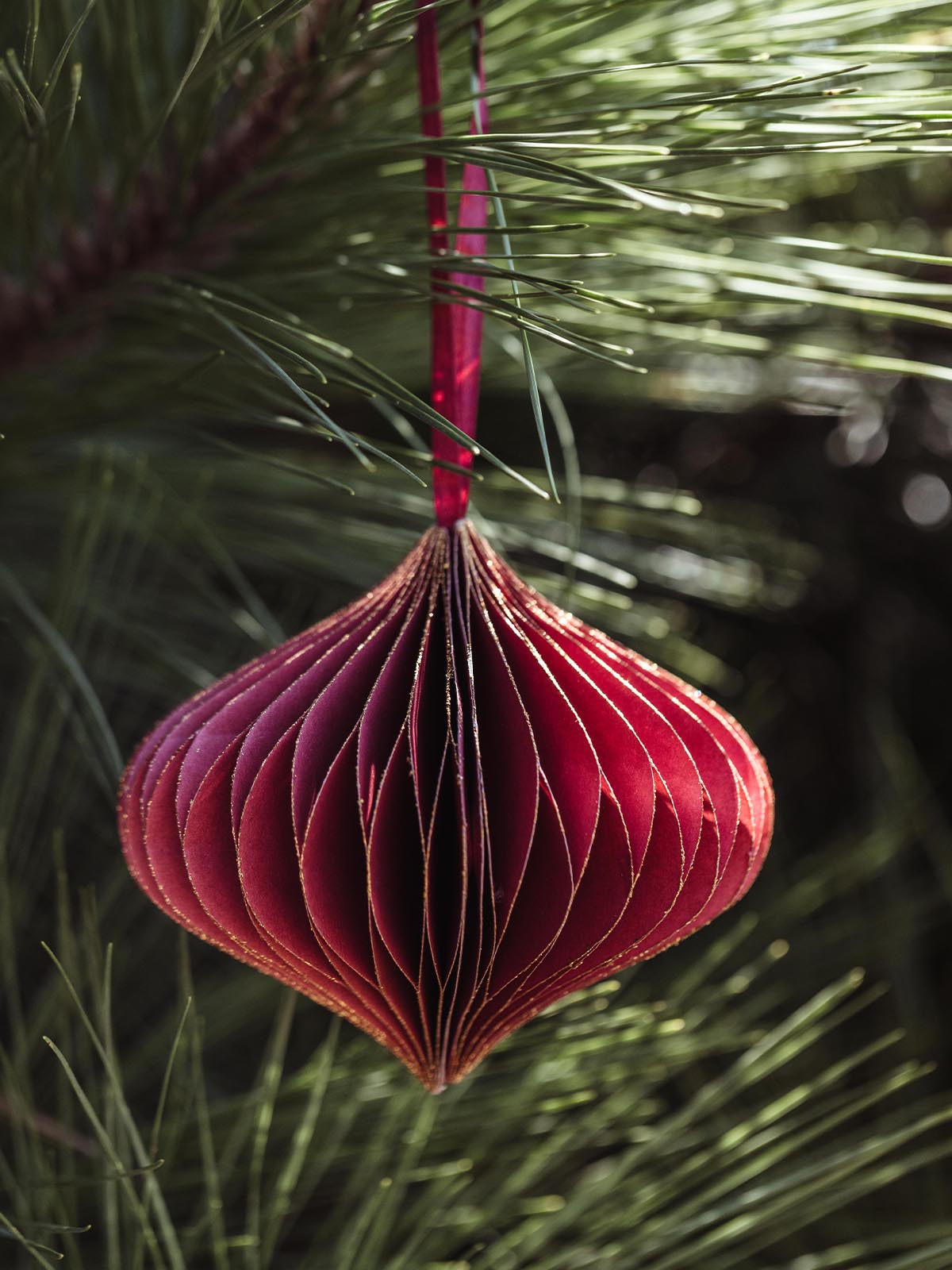 Single red handcrafted paper ornament with gold accents, hanging from a pine branch, offering a vibrant, festive touch to holiday decorations.