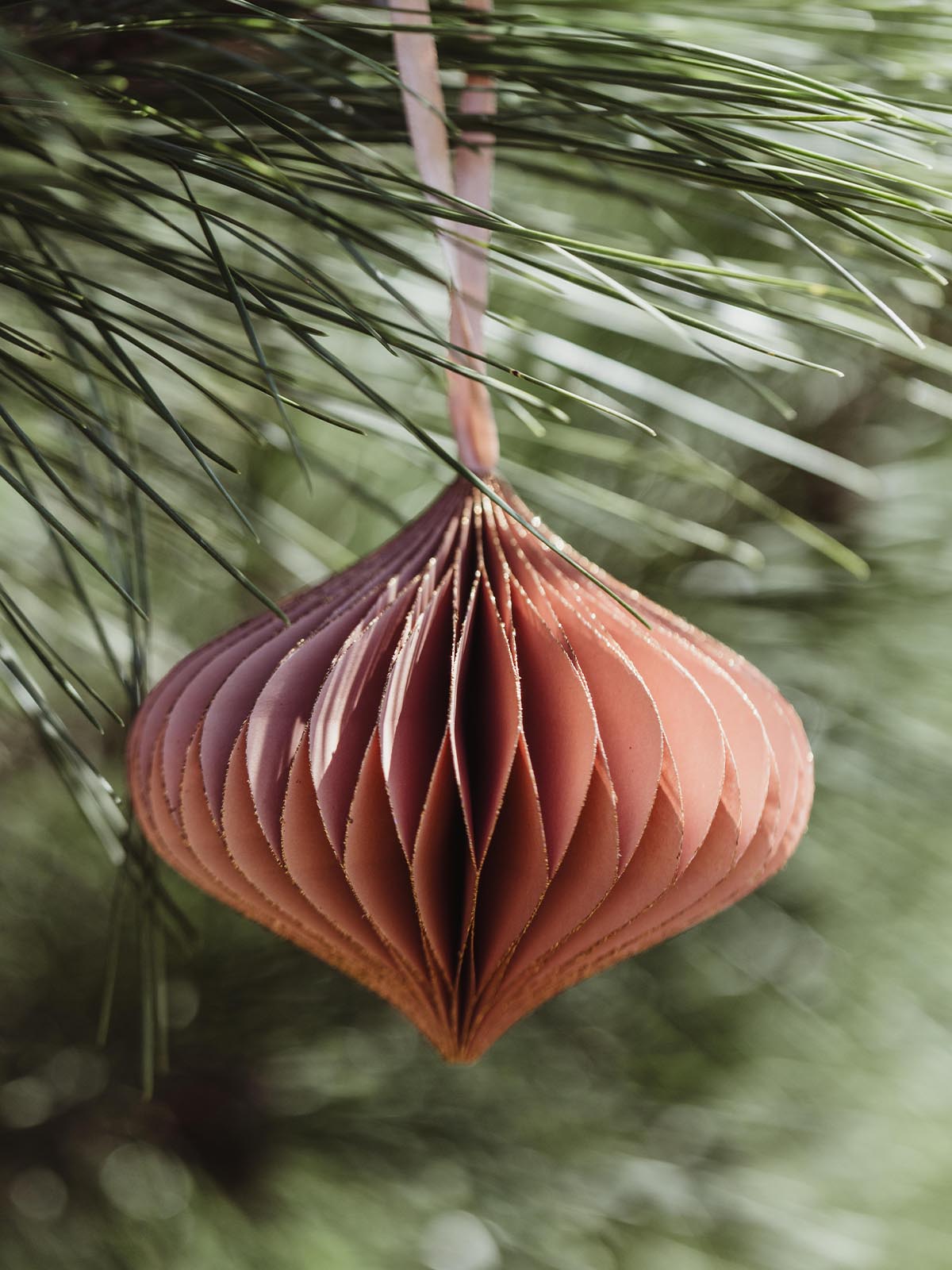 Single peach handcrafted paper ornament with gold accents, hanging from a pine branch, offering a vibrant, festive touch to holiday decorations.