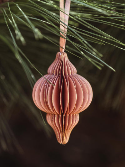 Elegant peach handcrafted paper ornament with detailed folds and a ribbon hanger, suspended from a pine branch, ideal for sustainable holiday decoration.