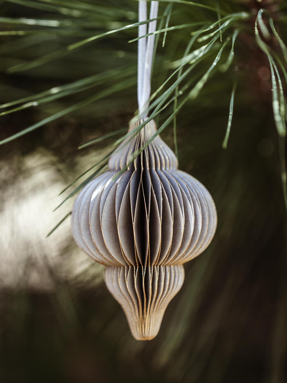 Elegant grey handcrafted paper ornament with detailed folds and a ribbon hanger, suspended from a pine branch, ideal for sustainable holiday decoration.