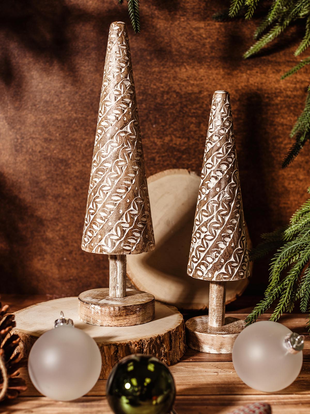 Hand-carved wooden Christmas tree decorations with intricate white patterns, standing on wood slice bases, surrounded by frosted glass and metallic holiday ornaments. Ethically made by artisans from Rahab's Rope.