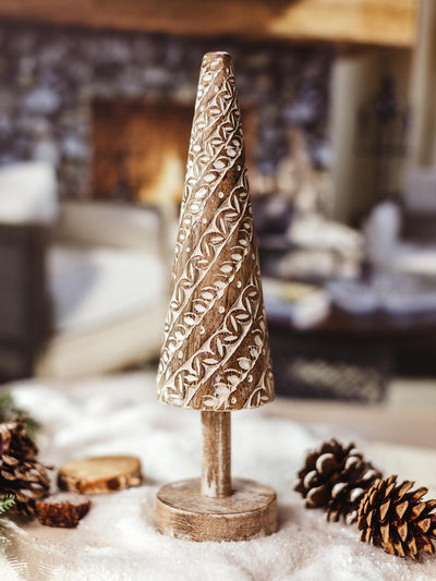 Hand-carved wooden Christmas tree decor with intricate white patterns, placed on a snowy surface with pinecones and a cozy fireplace in the background. 