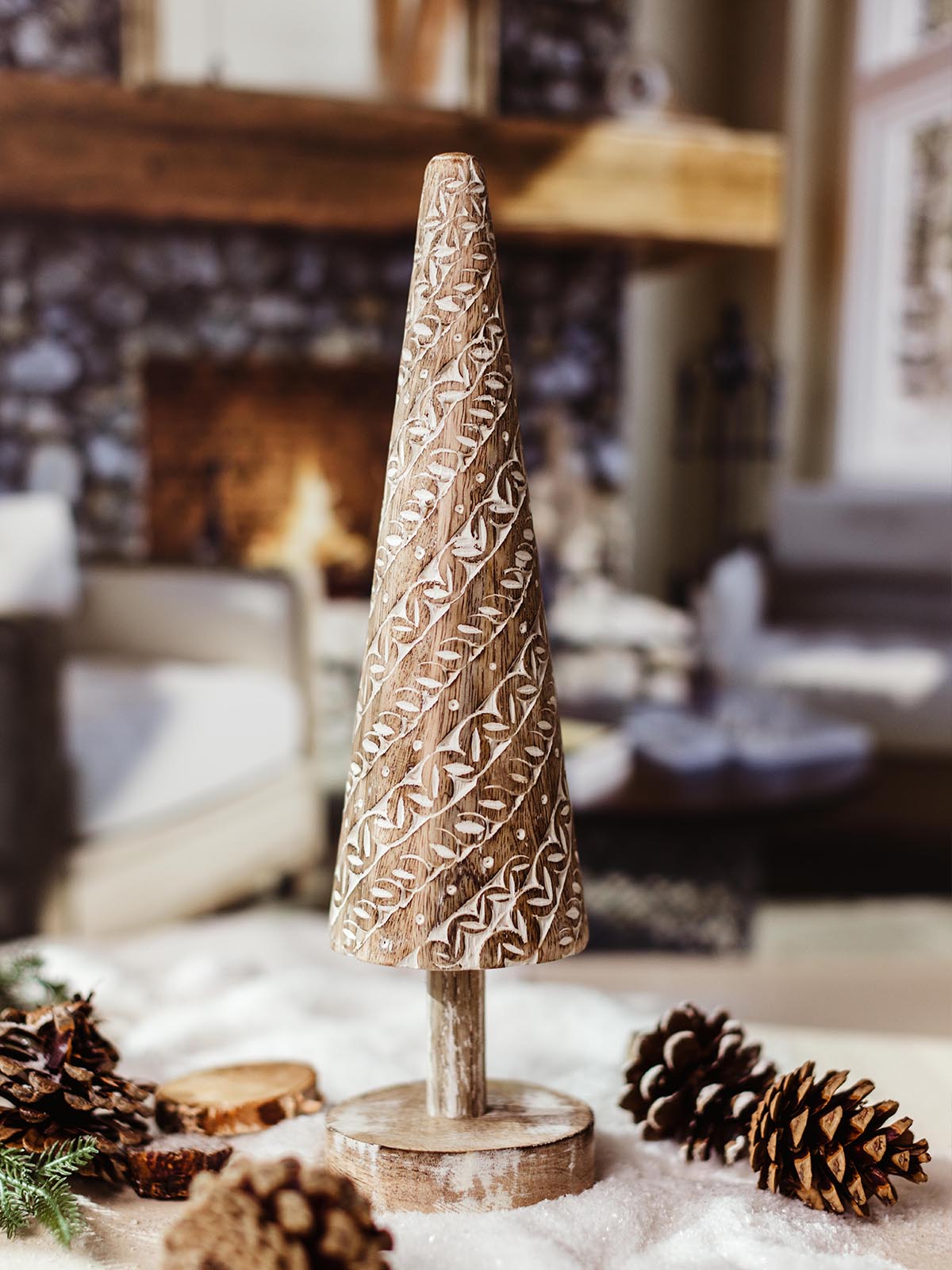 Hand-carved wooden Christmas tree decoration with white carved designs, standing on a wooden base amidst pinecones and snow. 