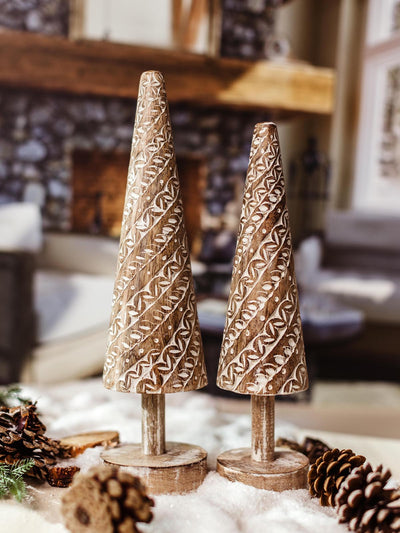 Two hand-carved wooden Christmas tree decorations featuring white swirl patterns, displayed on a snowy surface with pinecones and a fireplace setting. 