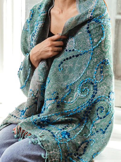 Woman wrapped in a green paisley shawl adorned with intricate blue embroidery and fringe, showcasing the detailed craftsmanship, part of Joffa.com's ethically sourced collection.