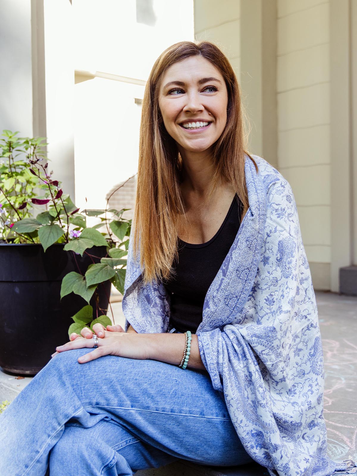 Smiling woman sitting outdoors wearing a soft blue and white paisley shawl, draped over her shoulders, part of Joffa.com's ethically handcrafted accessories collection.