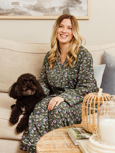 Smiling woman in a dark floral print pajama set sitting on a couch with a black dog, part of Joffa.com's ethically crafted loungewear collection.