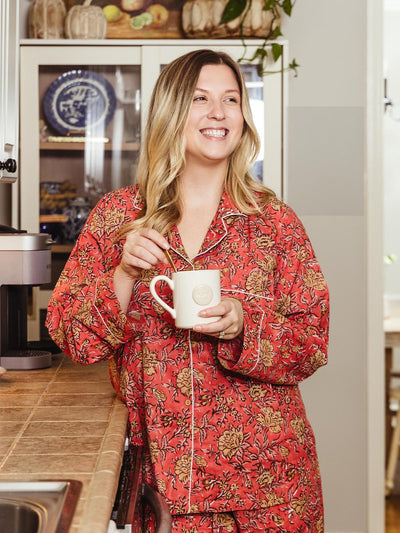 Woman smiling while holding a mug in the kitchen, dressed in a bright pink floral pajama set, showcasing Joffa.com's ethically made and cozy loungewear collection.