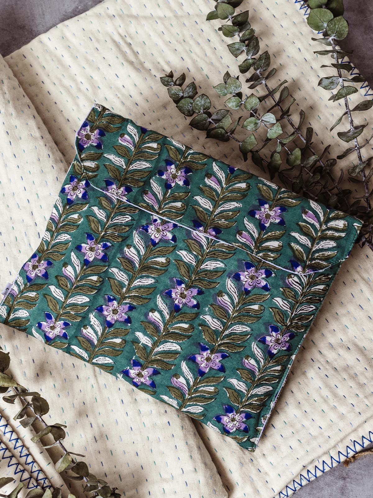 Ethically made green floral pouch featuring a purple flower and vine design, displayed on a cream quilt with blue stitching and accented with eucalyptus sprigs. This handcrafted pouch offers a beautiful and sustainable option for organizing personal items or gifting.