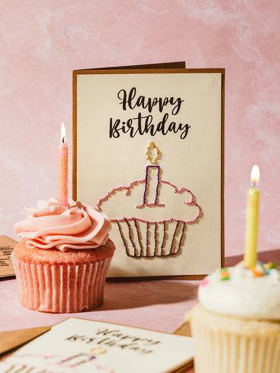 Handmade embroidered happy birthday card featuring a stitched cupcake design with a lit candle, displayed alongside pink and white frosted cupcakes with lit candles. Perfect for celebrating special occasions with a unique touch.