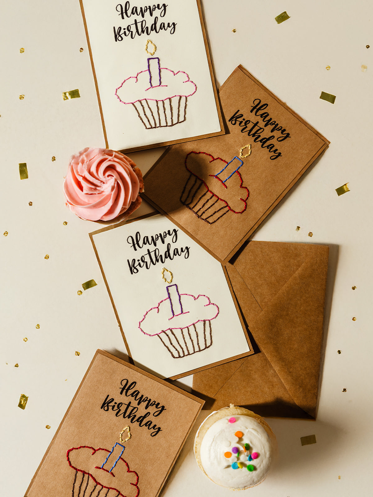 Flat lay of embroidered happy birthday cards with cupcake designs, surrounded by pink and white frosted cupcakes and gold confetti. A creative and handcrafted way to send birthday wishes.