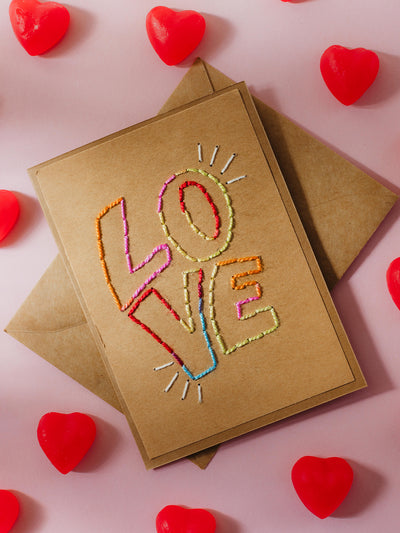 A kraft card featuring the word "LOVE" embroidered in vibrant rainbow thread, surrounded by small stitched details, paired with a matching kraft envelope. Red heart-shaped candies are scattered on a soft pink background.