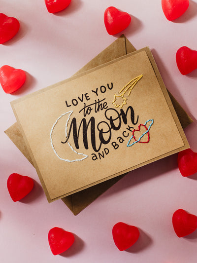  A kraft card with the phrase "Love you to the moon and back" in a mix of handwritten text and embroidery. It features a crescent moon, a shooting star, and colorful planetary rings. Red heart-shaped candies add a playful touch to the pink background.