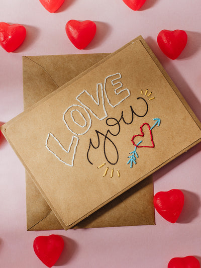 A kraft card with "LOVE you" written in black ink and white thread, featuring an embroidered red heart and arrow detail. A matching kraft envelope is included, with red heart-shaped candies scattered on the pink surface.