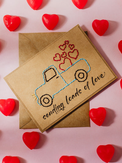  A kraft card with an embroidered vintage truck design carrying hearts and the text "sending loads of love." The card is surrounded by red heart-shaped candies on a light pink background.