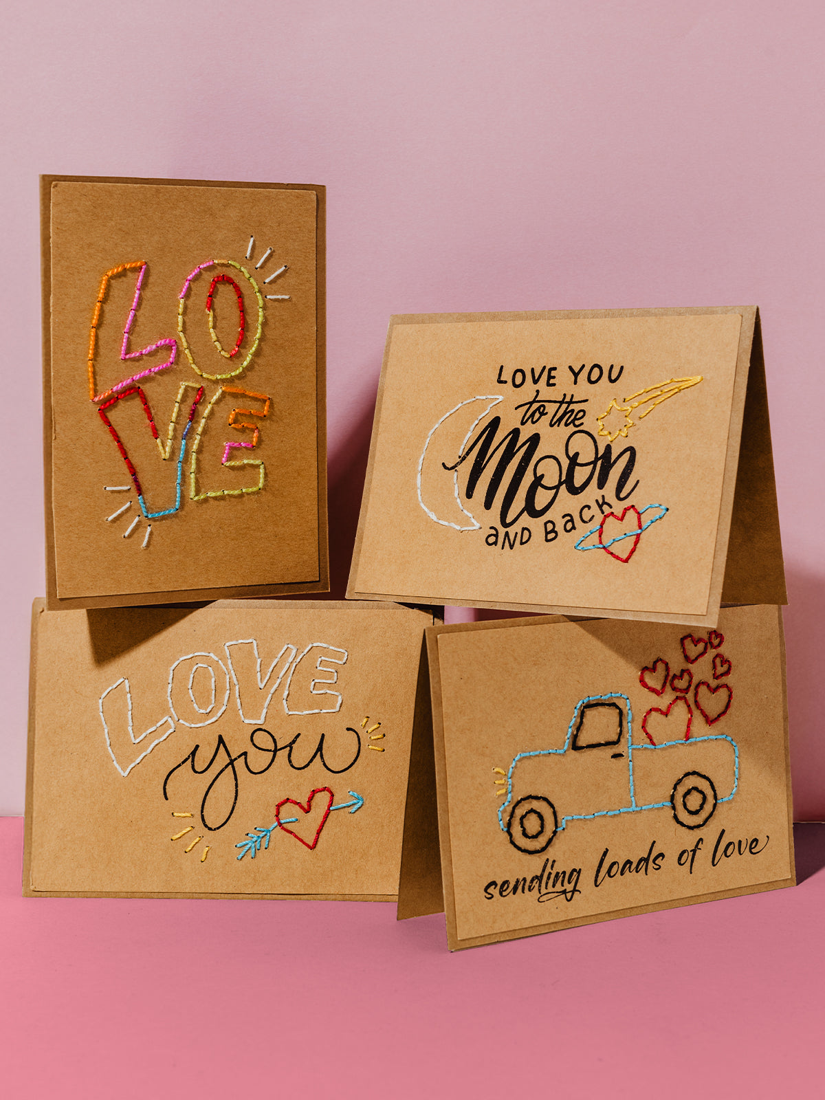  A collection of four kraft cards displayed together, showcasing various hand-embroidered designs: "LOVE" in rainbow colors, "Love you to the moon and back," "LOVE you" with a heart and arrow, and a truck carrying hearts with the phrase "sending loads of love." Set against a soft pink and kraft-toned backdrop.