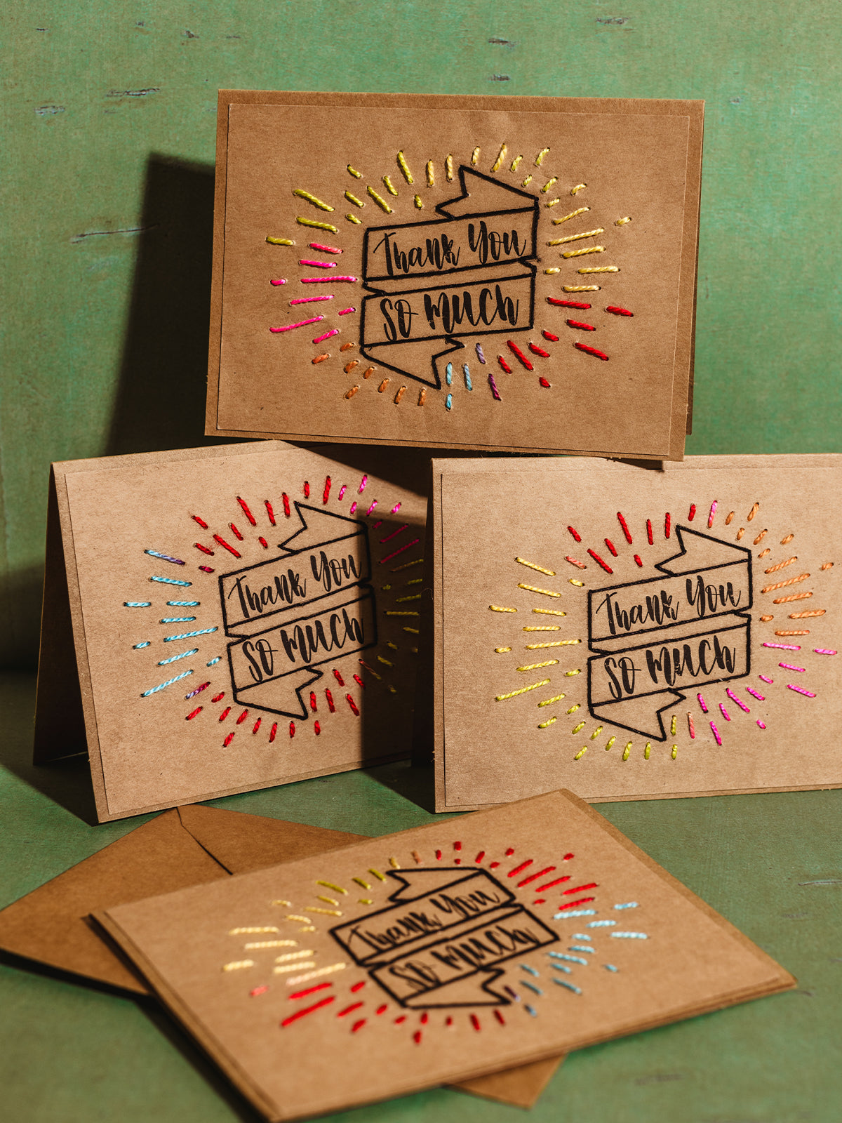 Handcrafted 'Thank You So Much' cards made from brown kraft paper with colorful embroidered accents, displayed on a green textured background, showcasing eco-friendly and artisan stationery perfect for thoughtful gifting.