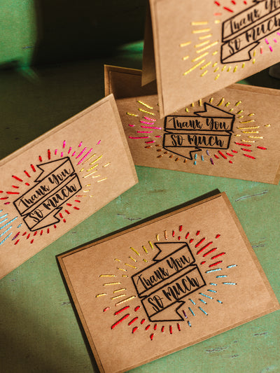 Handcrafted 'Thank You' cards made from brown kraft paper with colorful embroidered accents, displayed on a green textured surface, showcasing sustainable and artisan stationery design.