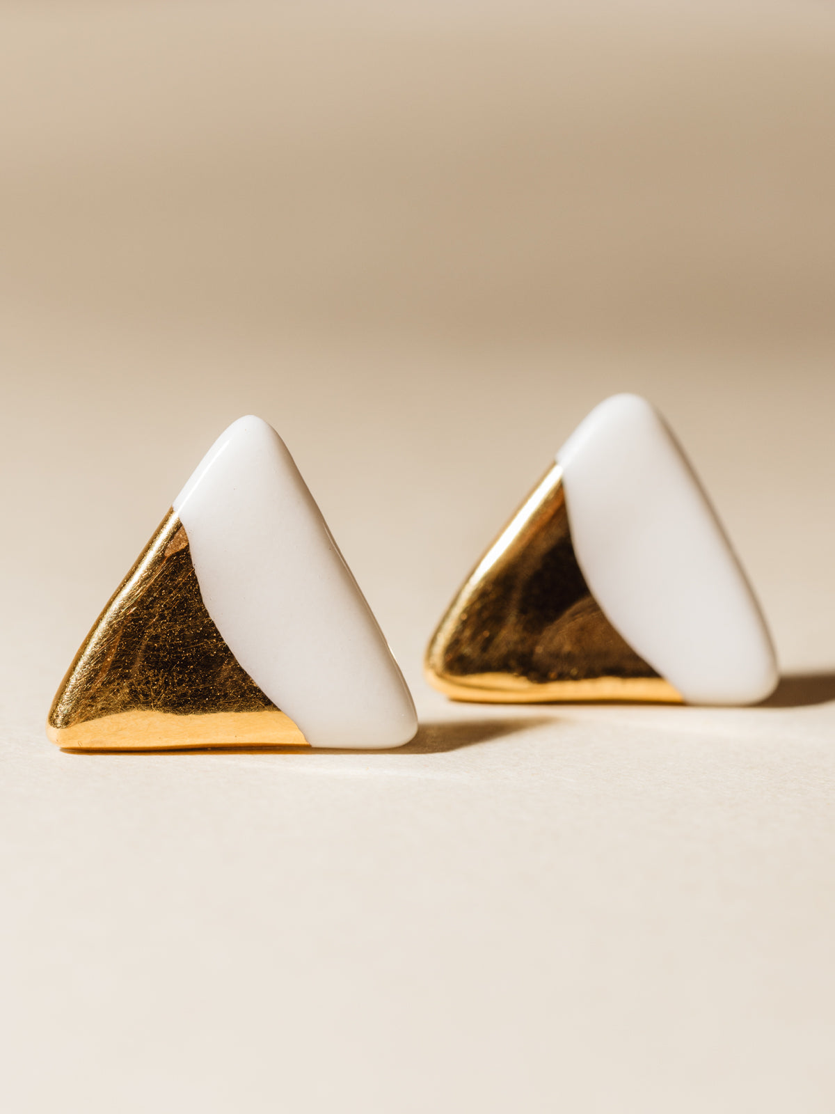 White triangle shaped clay stud earrings dipped in gold. Photographed on cream background.