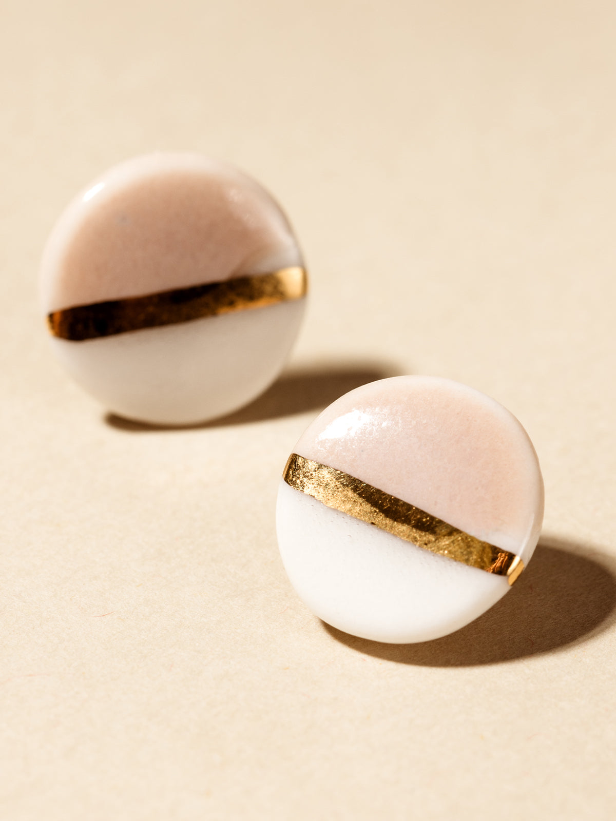 Circular clay stud earrings with white and blush color separated by gold fake line. Photographed on cream background. 