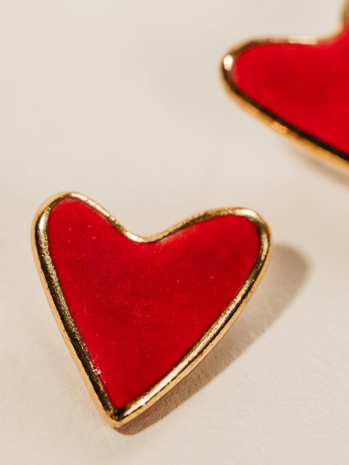 Close up of red heart clay stud earrings with gold outline. Photographed on cream background.