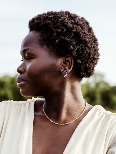 African female model wearing white dress. Model is also wearing gold chain necklace and porcelain diamond studs. Setting is sky and trees in the distance. 