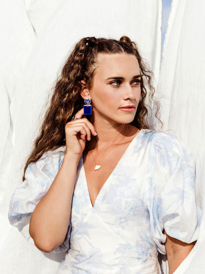 Portrait of white female model on white fabric background wearing light blue dress. Model is drawing attention to the blue flourish porcelain earring with her hand.