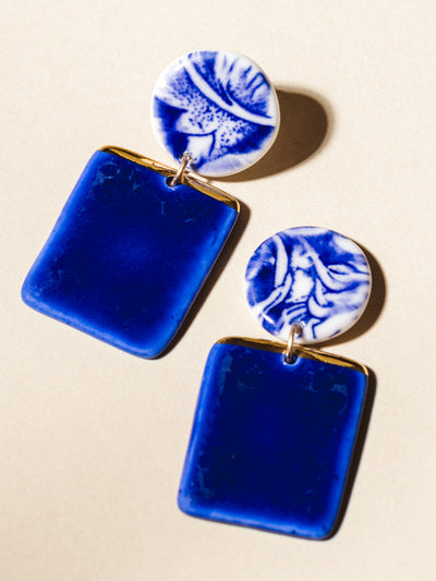 Vibrant blue ceramic earrings with circular stud and dangling rectangle shape dipped in gold. Photographed on cream background. 