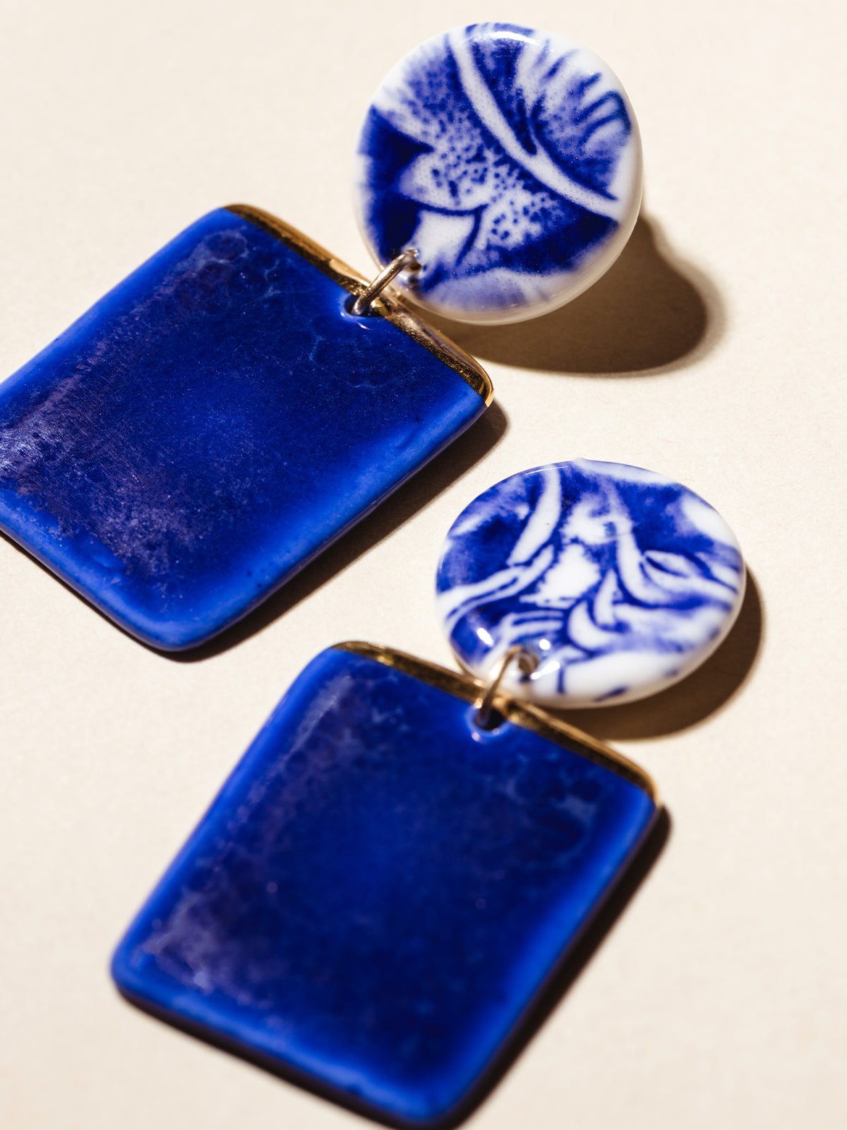 Vibrant blue ceramic earrings with circular stud and dangling rectangle shape dipped in gold. Photographed on cream background. 