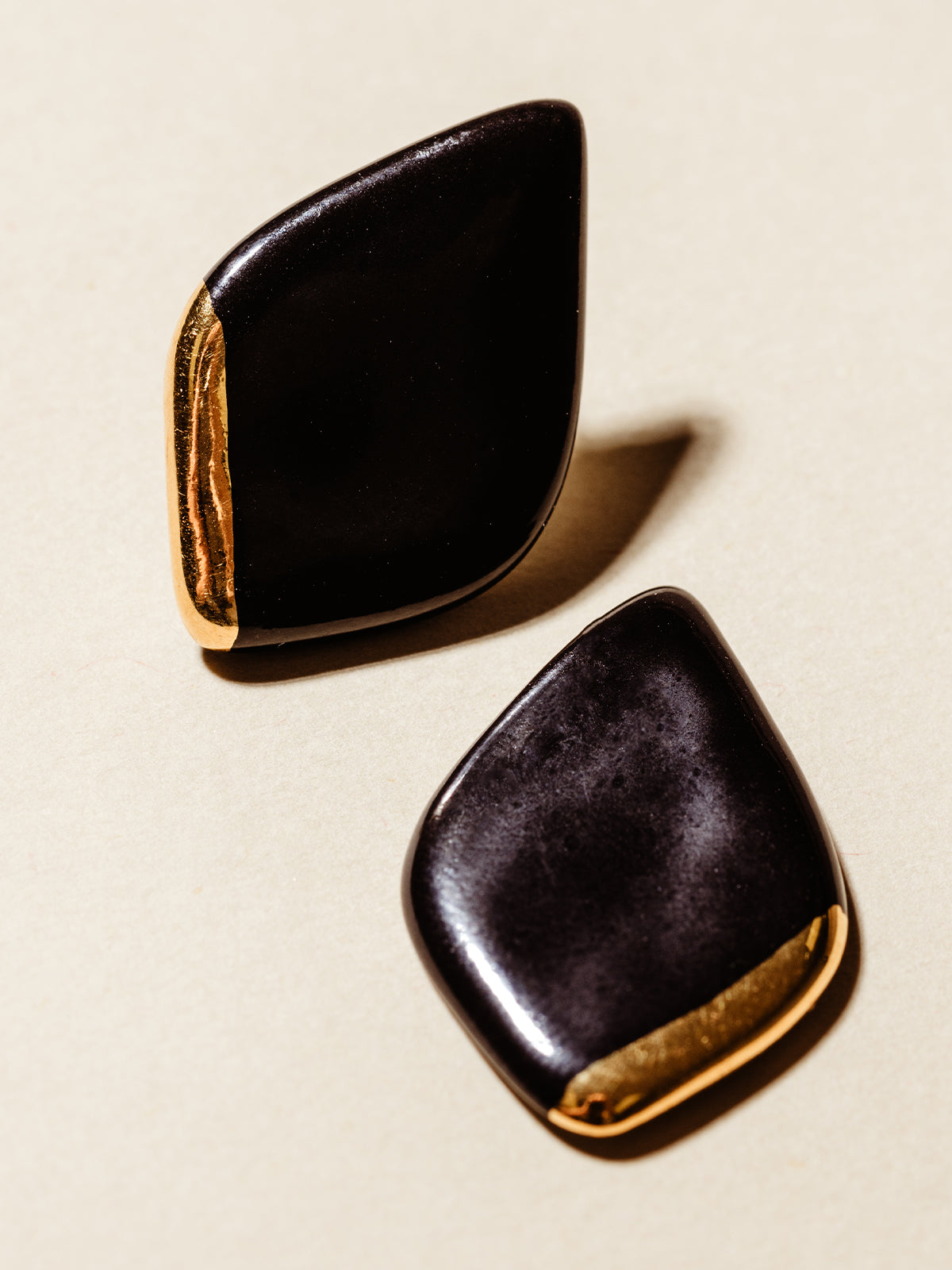 Black porcelain Dimond shaped earring studs with gold dipped edge. Photographed on tan background. 