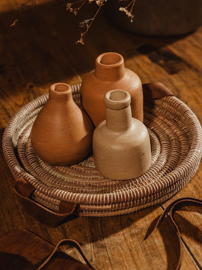 Round handwoven tray with cream center and cream threads. Tray is atop wooden table with clay vases placed inside tray on wood table.