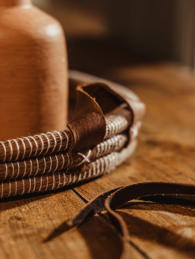 Close up of handwoven round tray leather handle and cream threads atop wood table surface.