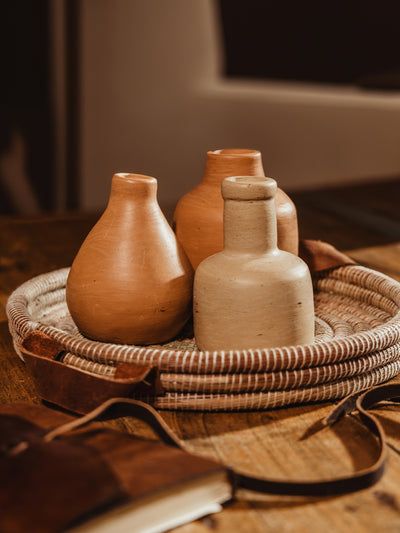 Round handwoven tray with cream center and cream threads. Tray is atop wooden table with clay vases placed inside tray on wood table.