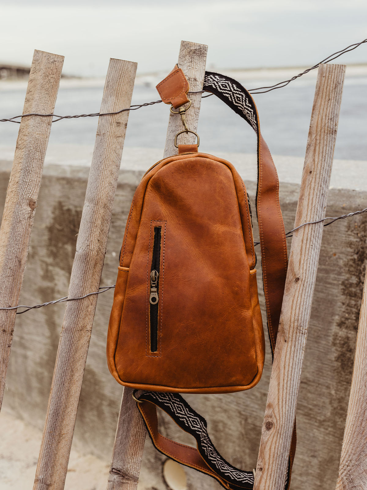 Handcrafted brown leather crossbody bag with a patterned strap hanging on a wooden fence by the beach, highlighting the bag’s rustic design and durability.