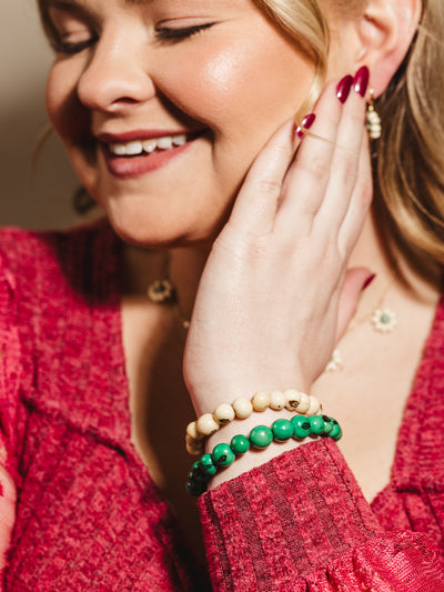 A smiling woman wearing a vibrant red sweater, accessorized with a stack of beaded bracelets featuring cream and green natural stone beads, styled with gold floral earrings and a matching necklace for a chic and bohemian look.