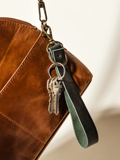 Dark green leather wristlet keychain with antique brass hardware, attached to a brown leather handbag. A stylish and practical accessory for secure key storage.