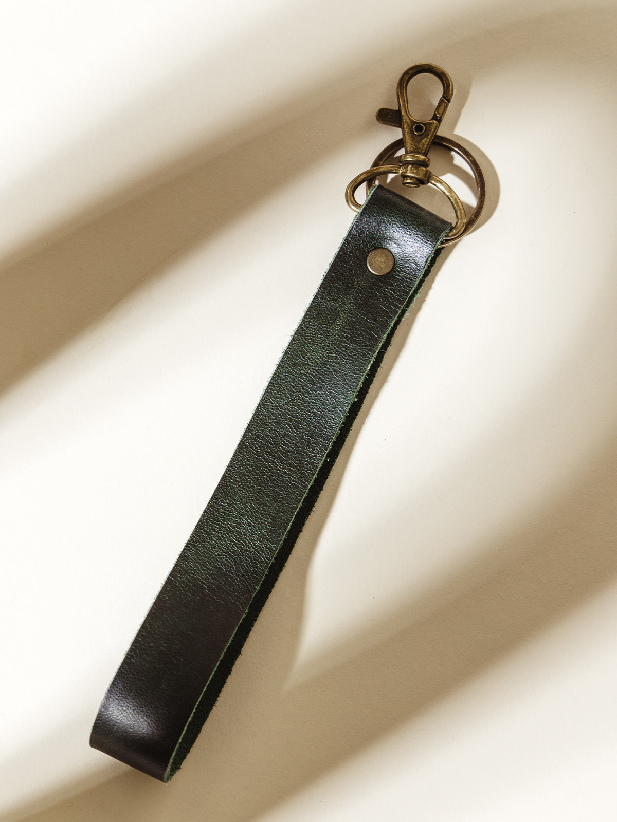 Close-up of a dark green leather wristlet keychain with brass clip and keyring, featuring a minimalist design. Perfect for organizing keys with an elegant touch.