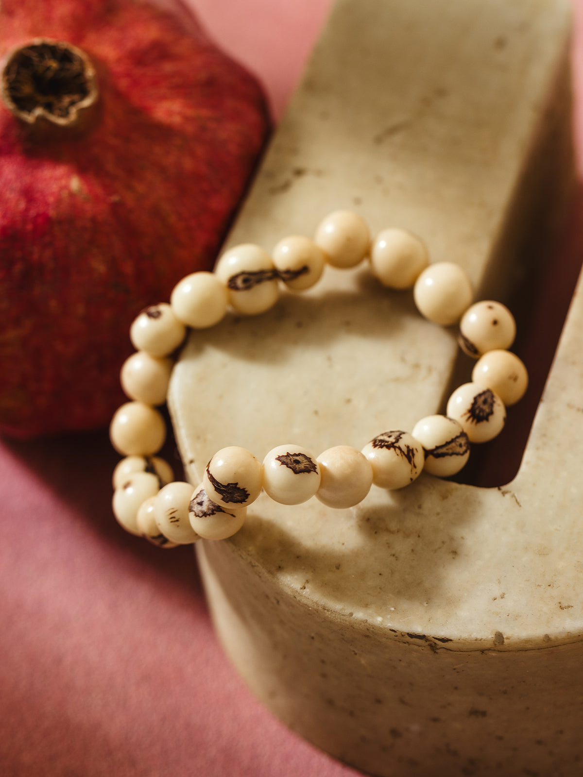 A close-up of a cream-colored beaded bracelet with dark brown natural markings on some beads, elegantly draped over a smooth, light beige stone surface. A red pomegranate with a rustic texture is visible in the background, adding a warm contrast to the neutral tones.