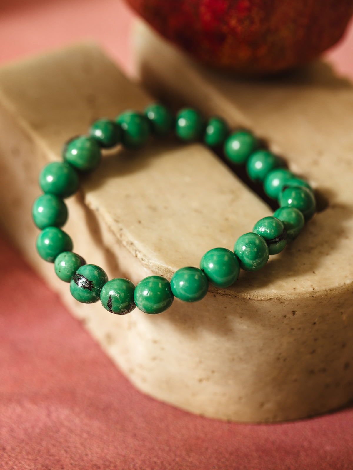 A vibrant green beaded bracelet with subtle black marbling details, displayed on a light beige stone surface. A red pomegranate is slightly blurred in the background, providing a rich contrast to the bright green beads and pinkish backdrop.