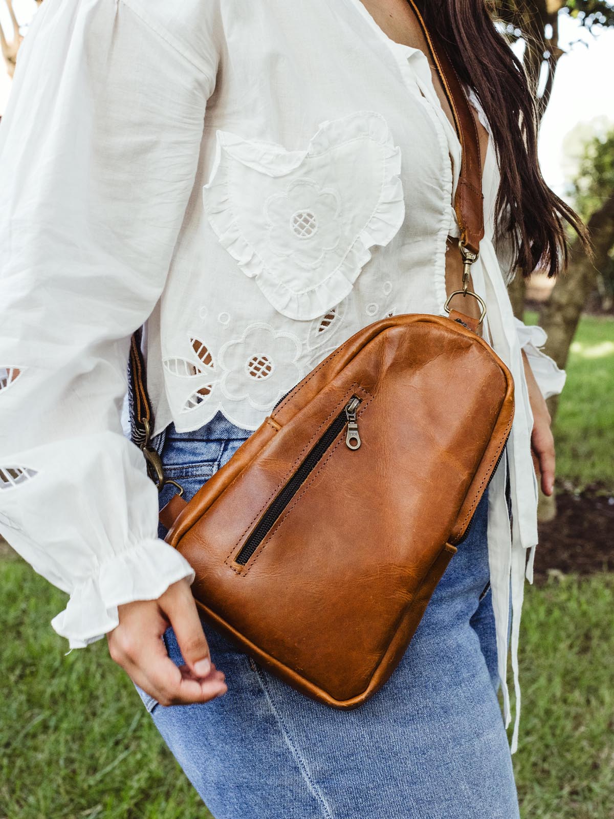 Side view of a woman wearing a brown leather crossbody bag, showcasing the bag’s design and zippered front pocket, paired with a white embroidered blouse and jeans.