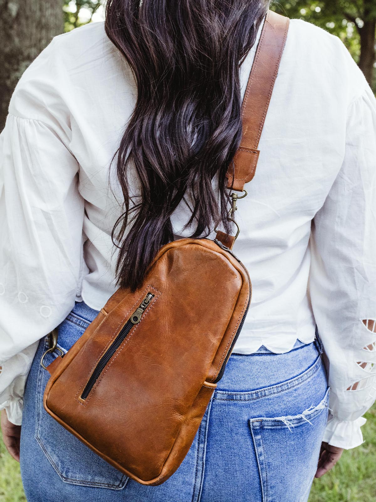 Back view of a woman wearing a handcrafted brown leather crossbody bag with a zippered front pocket, paired with a white blouse and jeans.